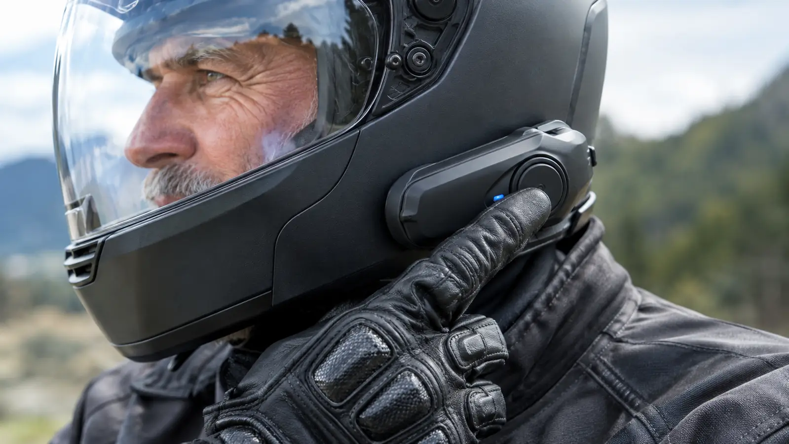 A rider's gloved hand pressing the main button on a helmet communicator mounted to the side of a helmet
