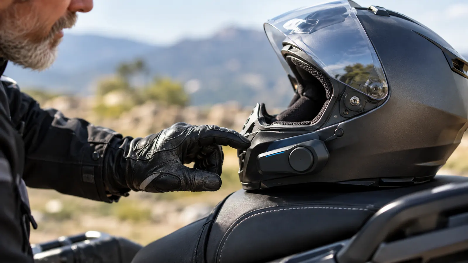 A rider's gloved hand reaching toward a small communicator mounted on the side of a helmet
