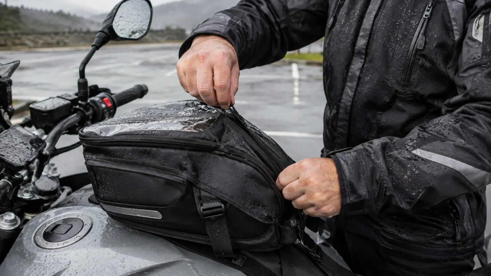 A rider zipping a rain jacket closed over a tank bag on a touring motorcycle