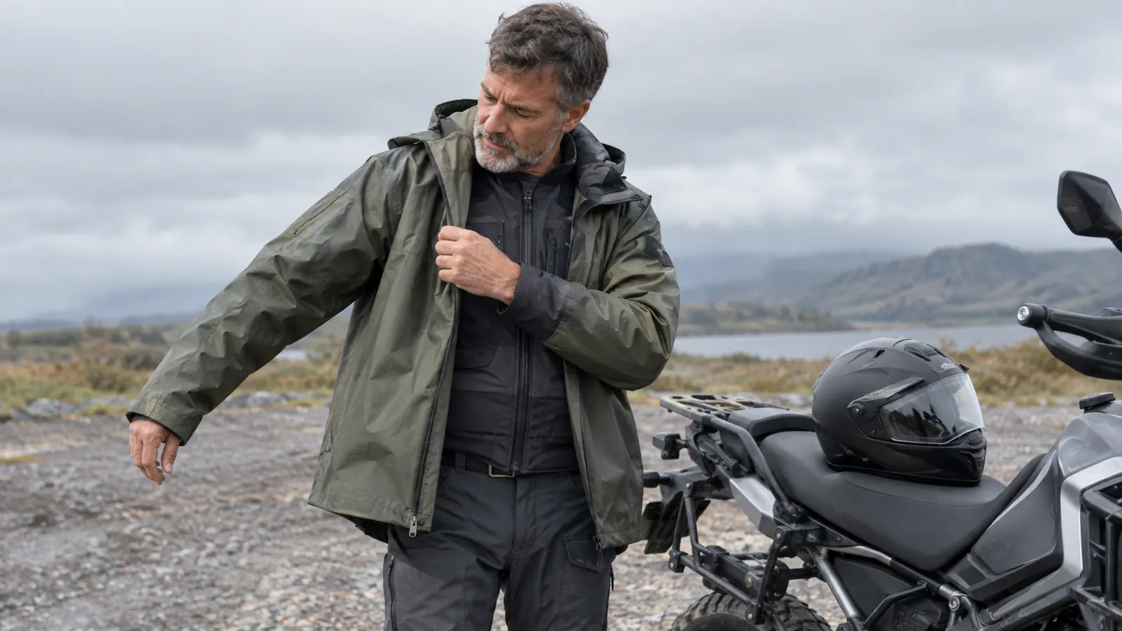 A rider pulling on the jacket of the rain suit over a riding jacket, parked in a gravel lot